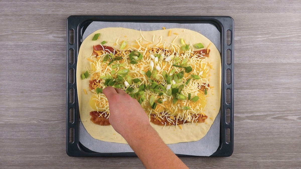 Käse und Frühlingszwiebeln werden über Pizzateig mit Kartoffeln und Bacon gestreut