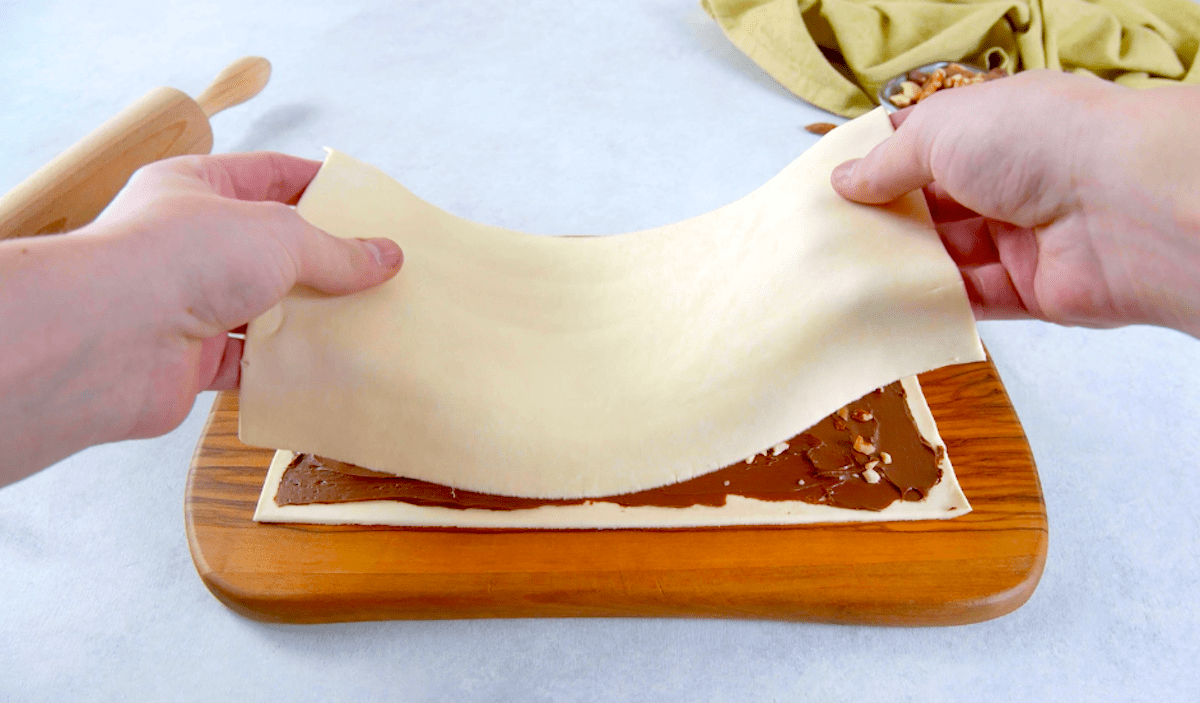 Ein Blätterteig wird auf einen anderen, bereits mit Nutella und gehackten Haselnüssen belegten Blätterteig gelegt.