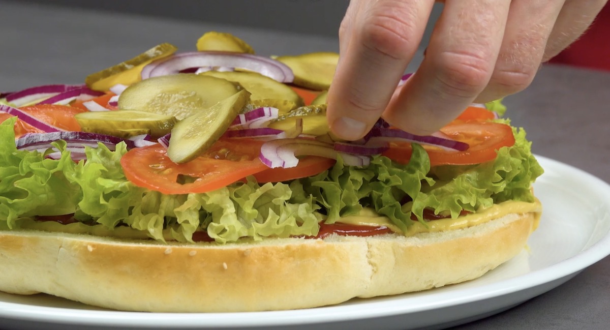 Untere Hälfte des Brötchens wird mit Salat, Gurken, Zwiebeln und Tomaten belegt