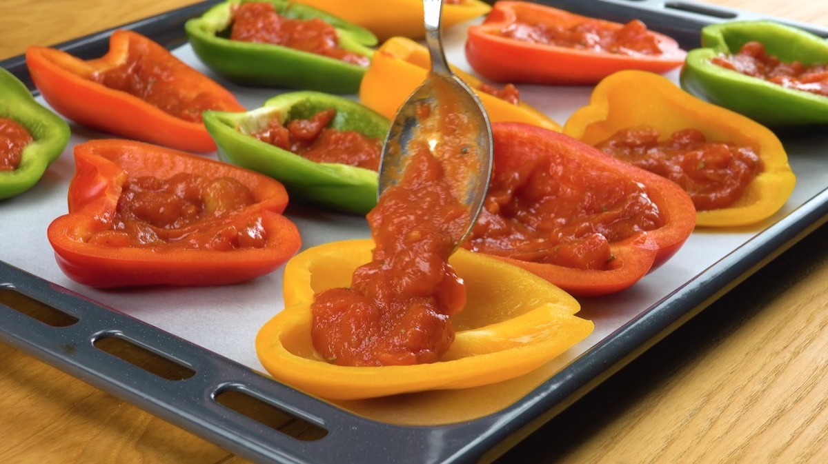 Halbierte Paprikas auf Backblech werden mit Tomatensoße gefüllt
