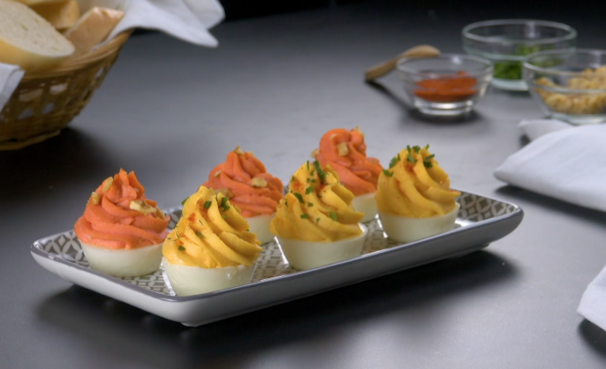 6 Teufelseier mit zwei unterschiedlichen Toppings auf einem länglichen Teller. Im Hintergrund ein Brotkorb und Gewürze in kleinen Schüsseln.