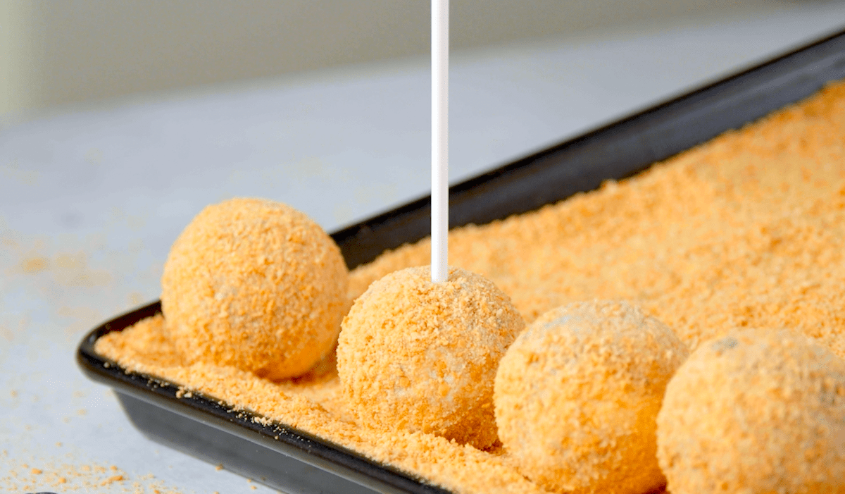 Ein Cake-Pop-Stiel wird in mit Kekskrümeln ummantelte Frischkäsebällchen gedrückt.