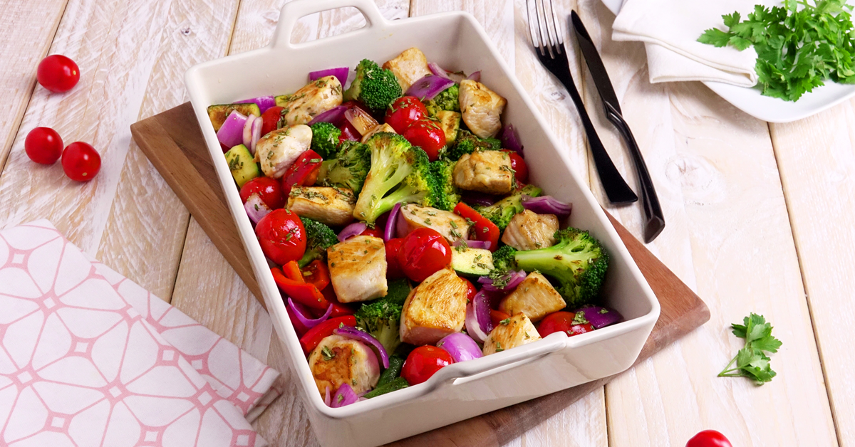 Hähnchen-Gemüse-Auflauf in Auflaufform. Daneben Besteck, Kräuter und kleine Tomaten.