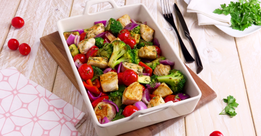 Hähnchen-Gemüse-Auflauf in Auflaufform. Daneben Besteck, Kräuter und kleine Tomaten.