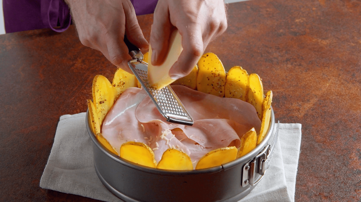 Eine Springform wird mit Kartoffelscheiben, Kochschinken und geriebenen Emmentaler gefüllt