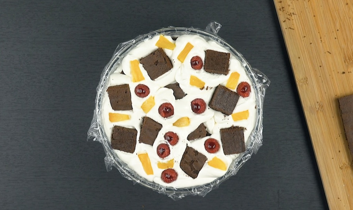 Mit Frischhaltefolie ausgekleidete Schüssel wird mit Frischkäsecreme gefüllt, in der Himbeeren, Ananasstücke und Kuchenquadrate verteilt werden