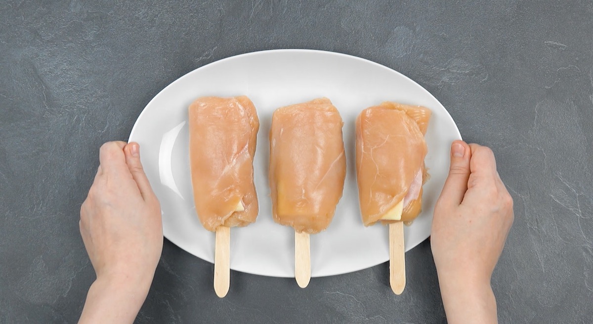 3 Hähnchenbrüste werden mit Kochschinken- und Käsescheibe belegt und um Holzstiel gewickelt