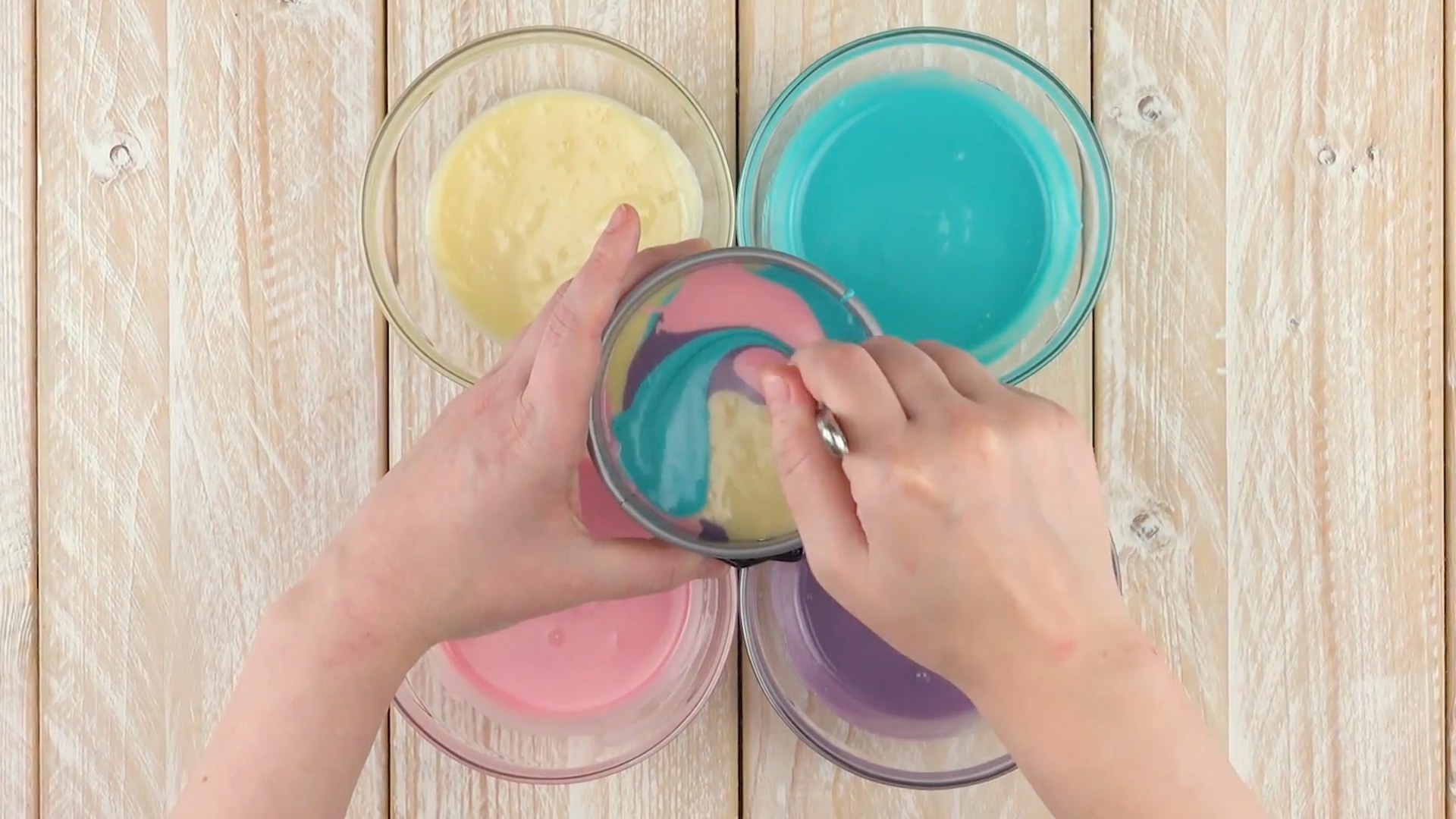 4 verschieden farbige Teige in Schüssel, die abwechselnd esslöffelweise in eine kleine Springform gegeben werden