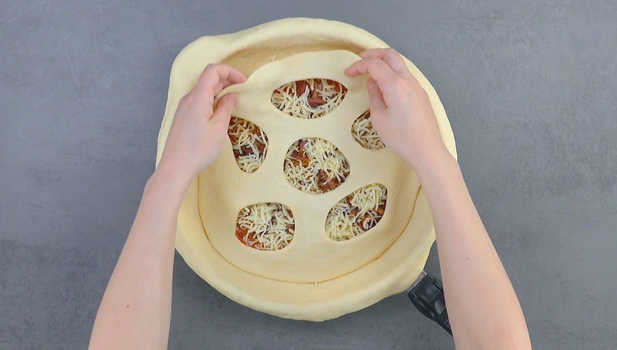 Gelöcherter Pizzateig wird auf Belag von Pfannenpizza gelegt