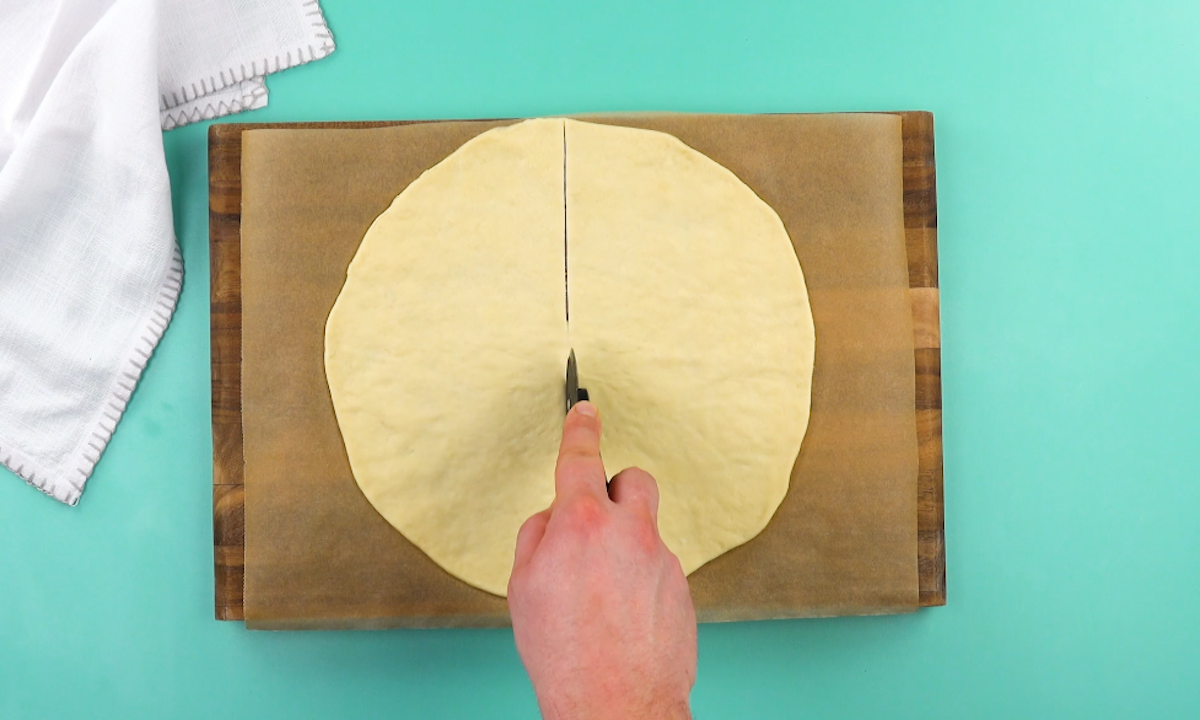 Kreisförmig ausgerollter Pizzateig wird mit Pizzaschneider halbiert.