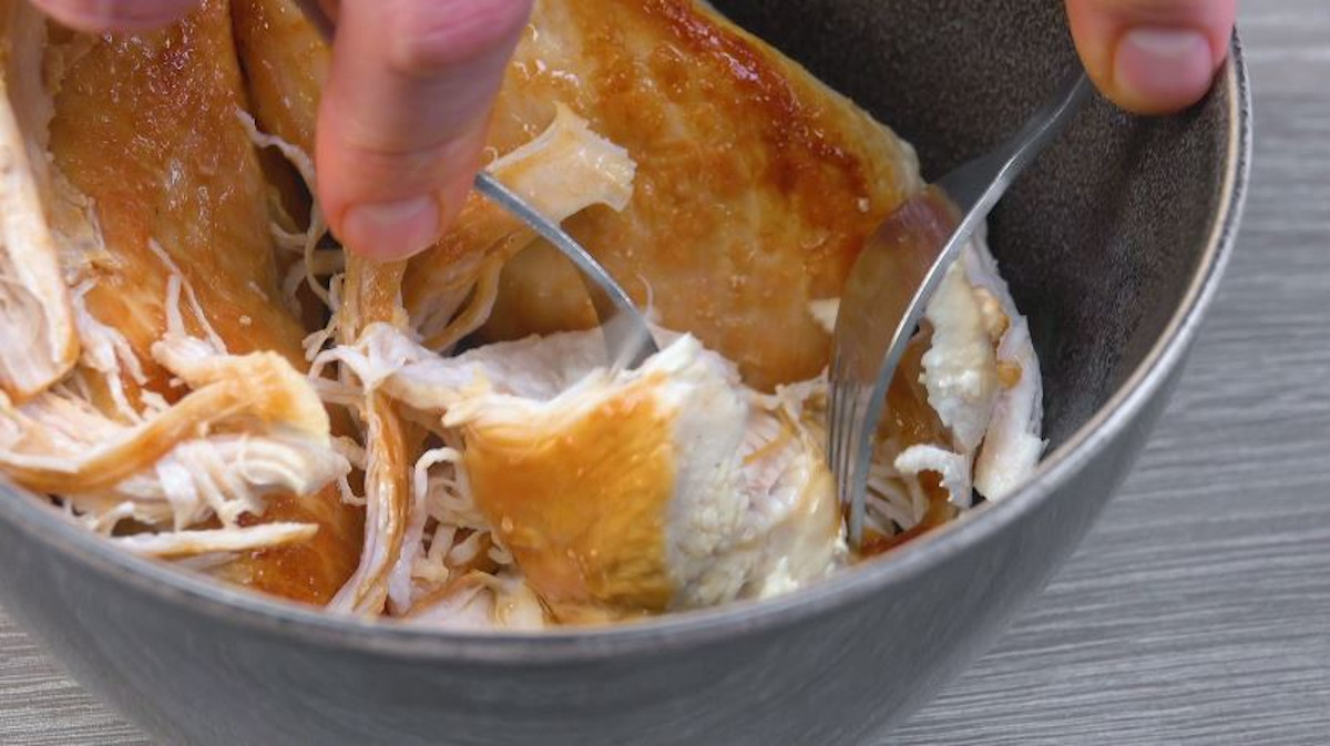 Angebratene Hähnchenbrüste werden mit Gabel in Schüssel auseinander gezogen