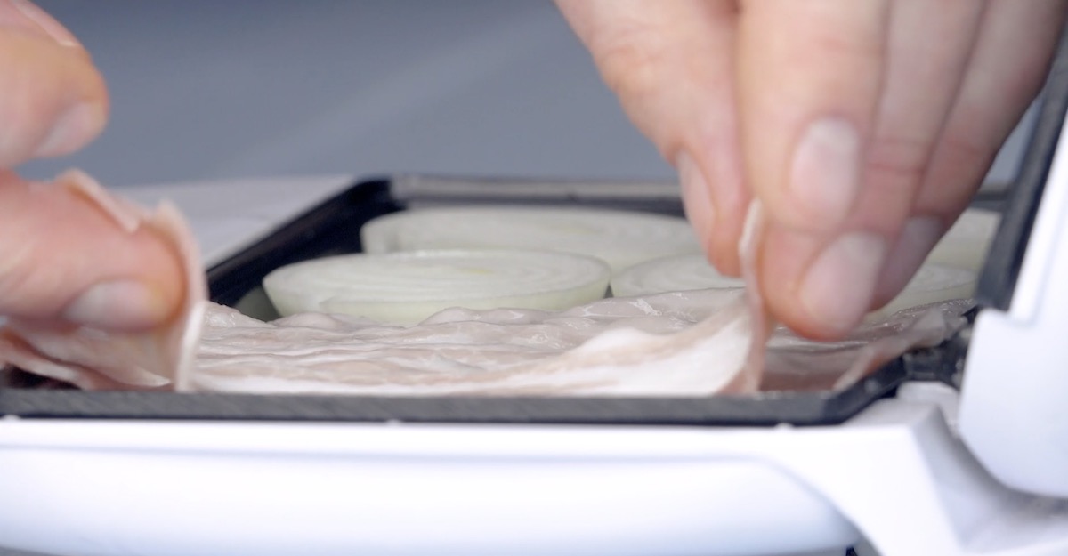 Bacon und Zwiebeln werden in geöffnetem Sandwichtoaster gebraten.