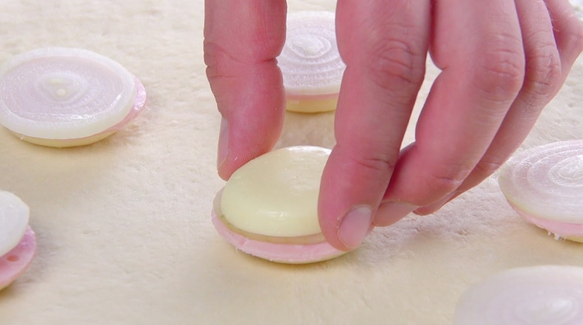 Halbierter Babybel-Käse wird auf Zwiebelring und Kochschinkenscheibe auf Pizzateig gelegt