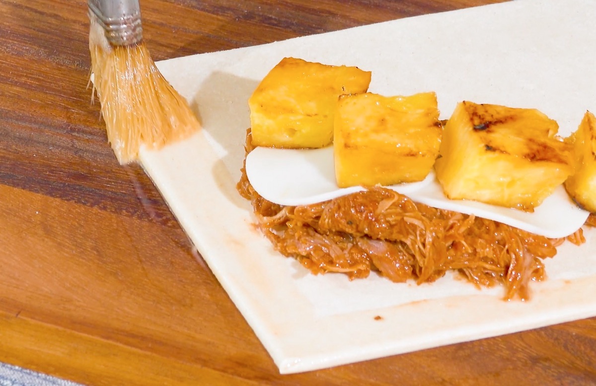 Blätterteig-Quadrat mit Ananas, Provolone und Pulled Pork wird am Rand mit Ei bepinselt