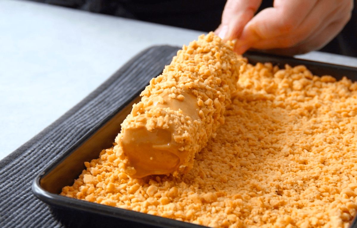 Aufgespießte Marshmallows, die mit Erdnussbutter ummantelt sind, werden durch klein gehackte Erdnüsse gedreht