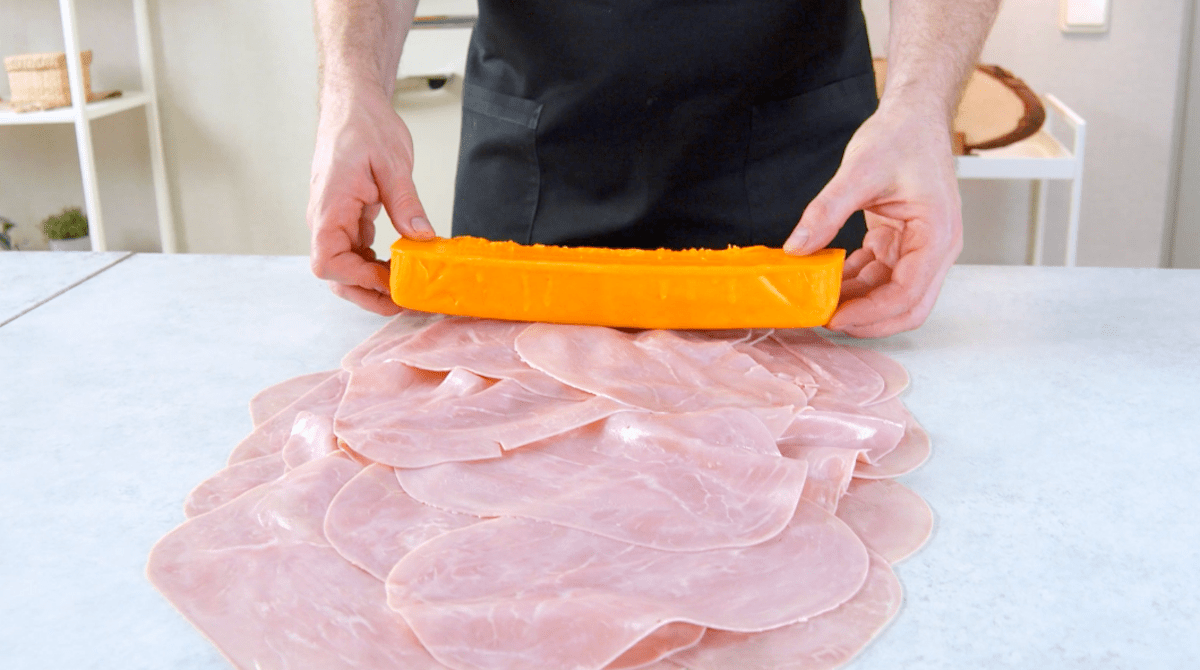 Ein Stück Cheddar wird auf mehrere Scheiben Kochschinken gelegt