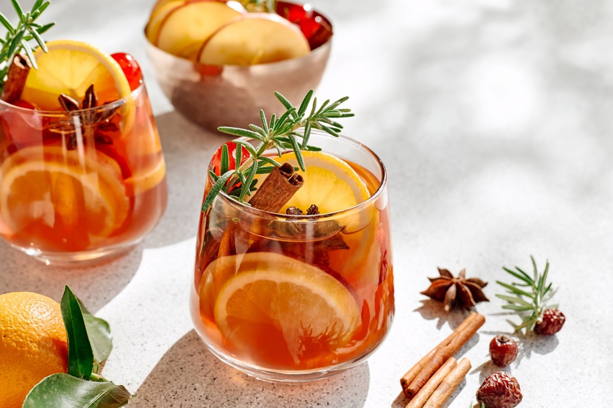 zwei Gläser weihnachtlicher Zitrus-Cocktail mit Zitrusscheiben, Zimtstangen, Sternanis und Rosmarin, daneben liegen Sternanis und Zimtstangen, im Hintergrund eine Schale mit Obstscheiben.