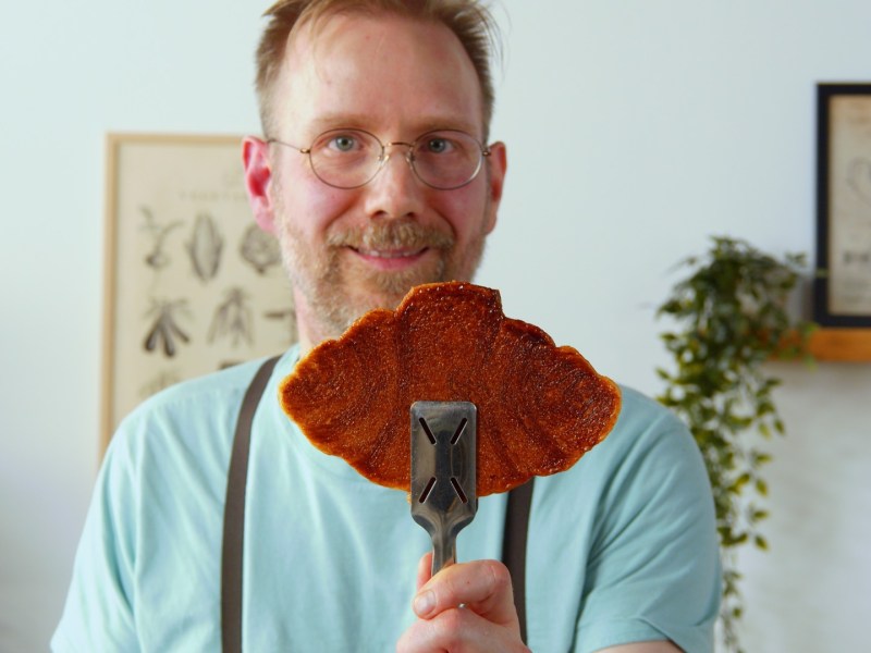 Ein Koch hält einen Croffles mit einer Zange in die Kamera.