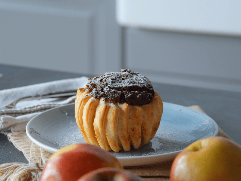 Blätterteig-Bratapfel mit Schokolade gefüllt auf einem Teller, im Vordergrund unscharf ein paar Äpfel.