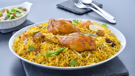 Ein tiefer Teller mit Chicken Biryani. Im hintergrund sind Besteck und eine Schale mit Salat.