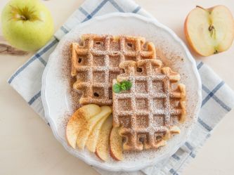 Haferflocken-Apfelmus-Waffeln angerichtet auf einem Teller mit Apfelspalten und Puderzucker. Der Teller steht auf einem Küchenhandtuch. Zwei Äpfel liegen als Dekoration neben dem Teller.