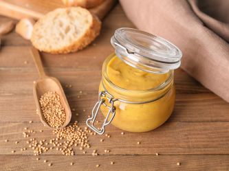 Honig-Senf-Soße in einem Bügelglas serviert. Senfkörner, Weißbrot und eine Serviette liegen im Hintergrund.