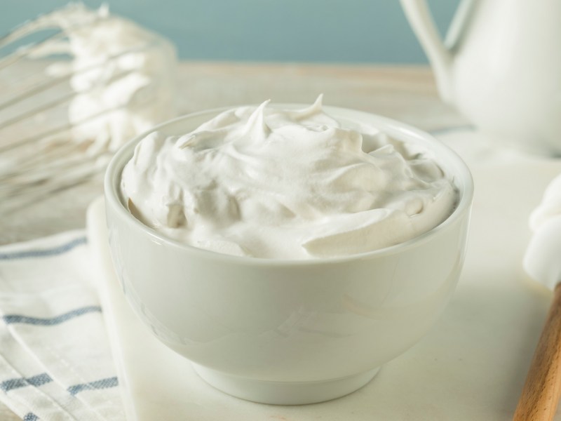 Rahmprodukte im Vergleich: süße Schlagsahne in einer Schüssel aufgeschlagen, daneben ein Rührbesen und ein Backspatel.