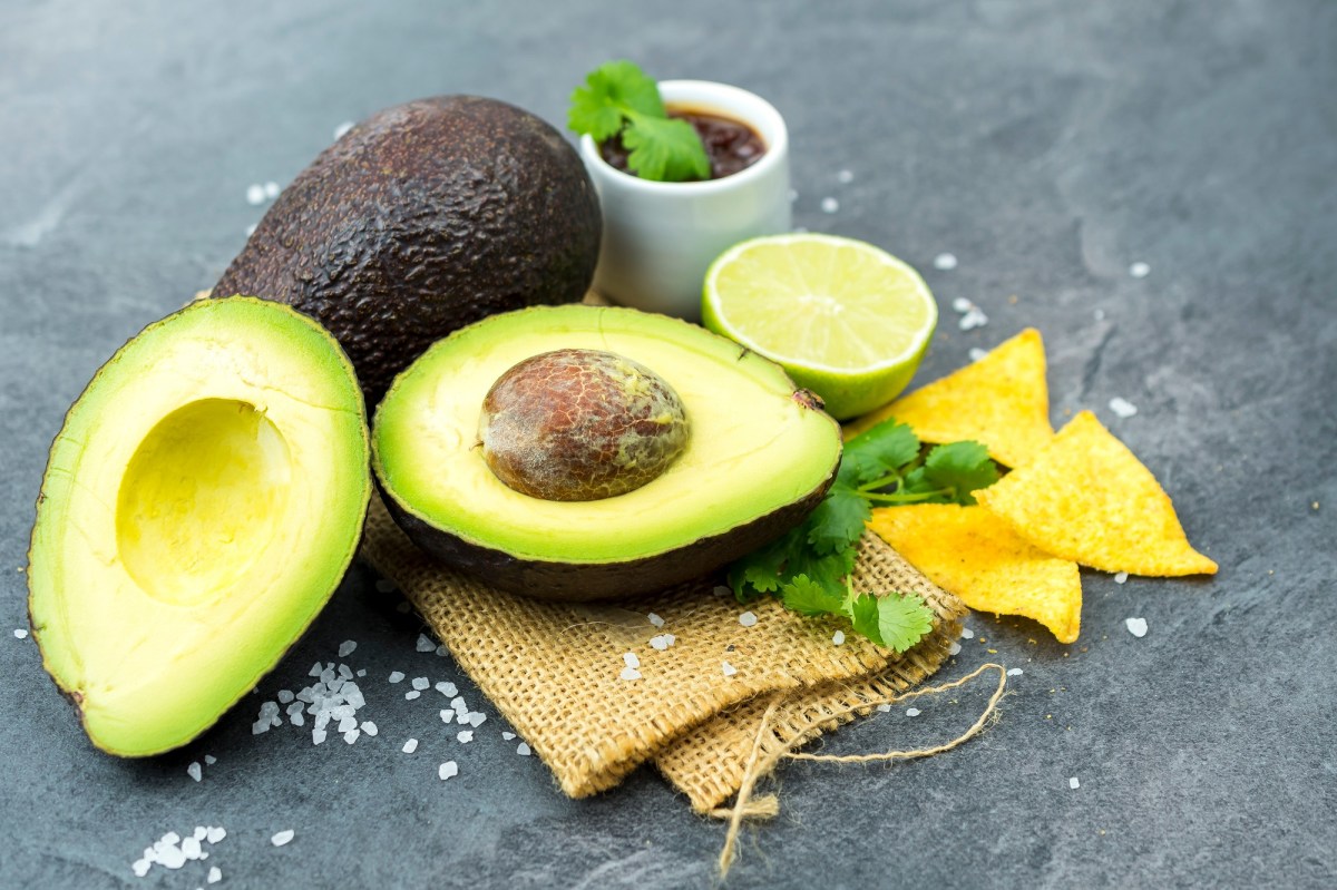 Avocado schneller reifen lassen: Eine ganze und eine halbierte Avocado, daneben Nachos, eine halbe Limette und ein Schälchen mit Dip.