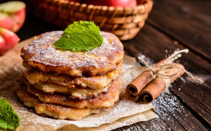 Ein Stapel Apfelpfannkuchen mit Puderzucker. Im Hintergrund sind frische Äpfel zu sehen, daneben liegen Zimtstangen.