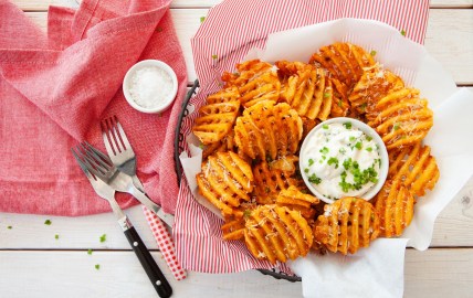 Gitterkartoffeln auf einem Teller, in der Mitte eine Schale weißer Dip, daneben Besteck und ein Geschirrtuch, Draufsicht.
