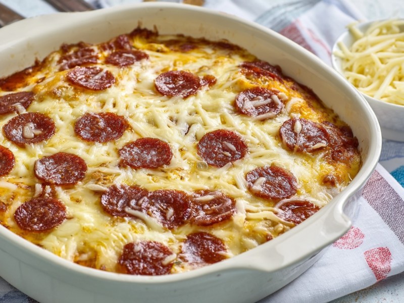 Pizza-Auflauf in weißer Auflaufform daneben eine Schale mit geriebenem Käse.