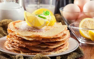 Ein Stapel britische Pancakes mit Zitronenstücken obendrauf. Im Hintergrund stehen die Zutaten.