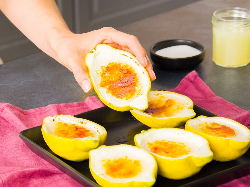 Ein kleines Blech mit sechs mal Lemon Posset, das eine karamellisierte Kruste hat. Köchin Adrianna hält eine Zitronenhälfte. Im Hintergrund kann man eine Schale mit Zucker und ein kleines Glas mit Zitronensaft sehen. Das Blech mit den Zitronen steht auf einem dunkelpinken Küchentuch.