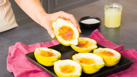 Ein kleines Blech mit sechs mal Lemon Posset, das eine karamellisierte Kruste hat. Köchin Adrianna hält eine Zitronenhälfte. Im Hintergrund kann man eine Schale mit Zucker und ein kleines Glas mit Zitronensaft sehen. Das Blech mit den Zitronen steht auf einem dunkelpinken Küchentuch.