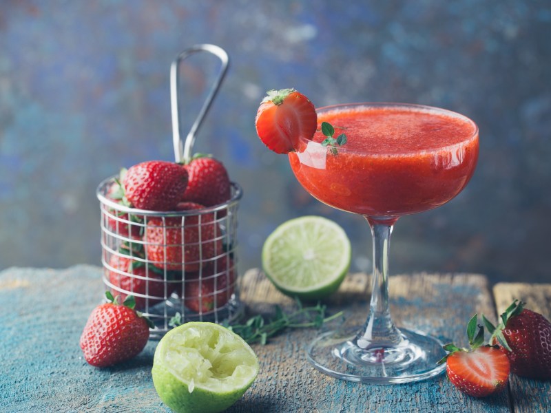 Erdbeer-Daiquiri in einem Cocktailglas, garniert mit Erdbeere und Thymian. Daneben liegen Limetten und weitere Zutaten.