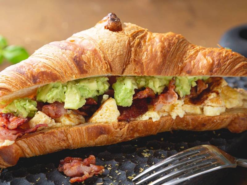 Ein Croissant mit Rührei Avocado und Speck auf einer Schieferplatte, davor eine Gabel.