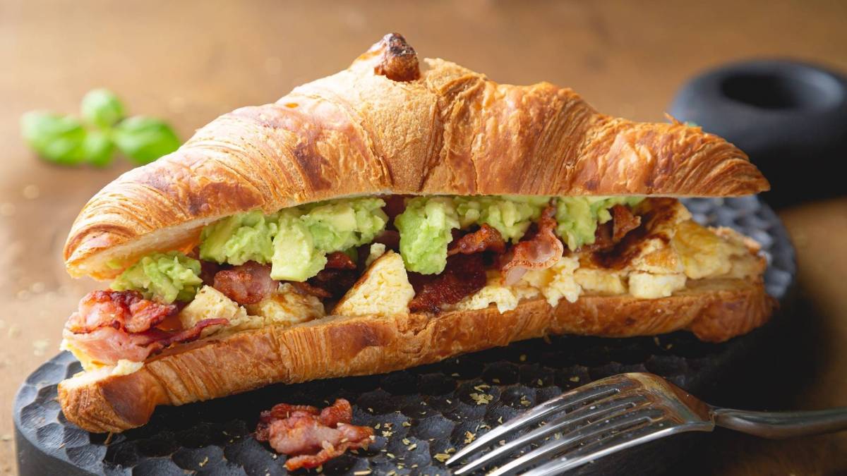 Ein Croissant mit Rührei Avocado und Speck auf einer Schieferplatte, davor eine Gabel.