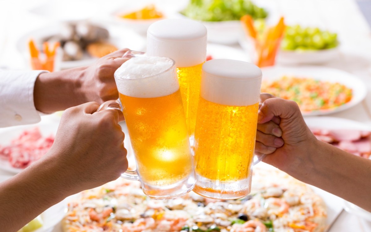 Tag des Biers: 3 Bierkrüge werden aneinander gestoßen. Im Hintergrund ist Essen zu sehen.