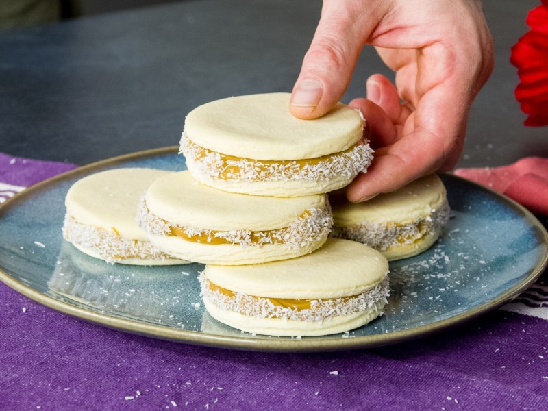 Ein Stapel Alfajores auf einem Teller. Jemand nimmt sich den obersten Keks.