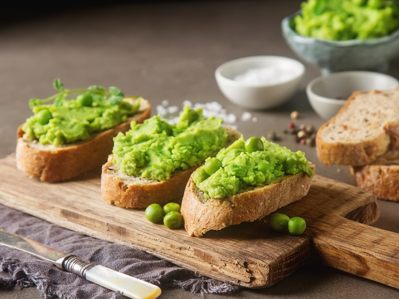 Erbsen-Basilikum-Hummus auf 3 Baguettescheiben, die auf einem Brett liegen. Im Hintergrund eine Schüssel mit dem Hummus und Brotscheiben.