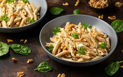 Ein grauer Teller mit einer großen Portion Hummus-Pasta, die mit frischer Petersilie und gehackten Walnüssen serviert wird. Im Hintergrund ist eine weitere Portion des Gerichts zu sehen. Auf dem Tisch liegen Basilikumblätter und Walnussstückchen verteilt.