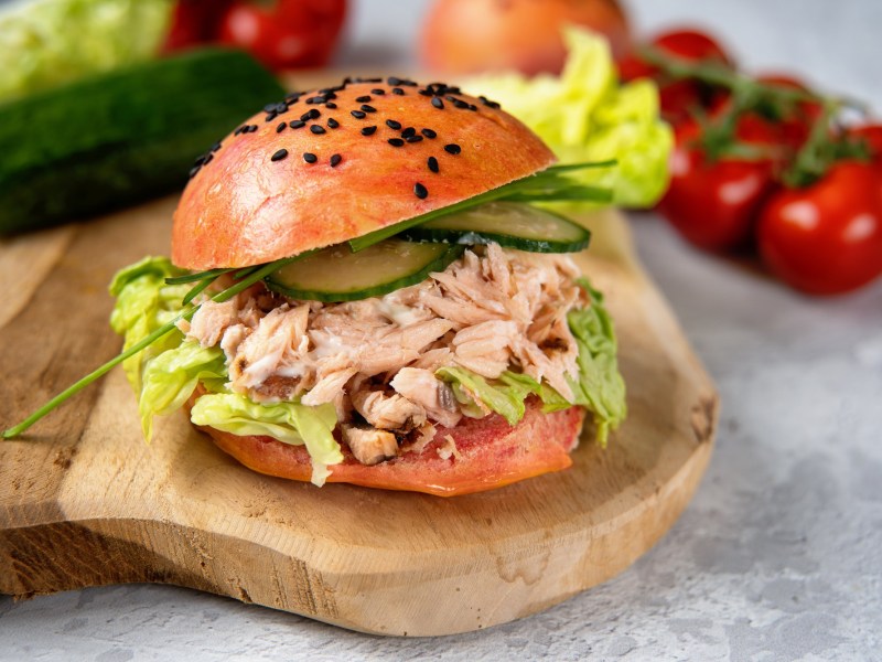 Ein Lachs-Burger mit Gurken und Salat auf einem Brett, im Hintergrund frische Zutaten wie Tomaten.