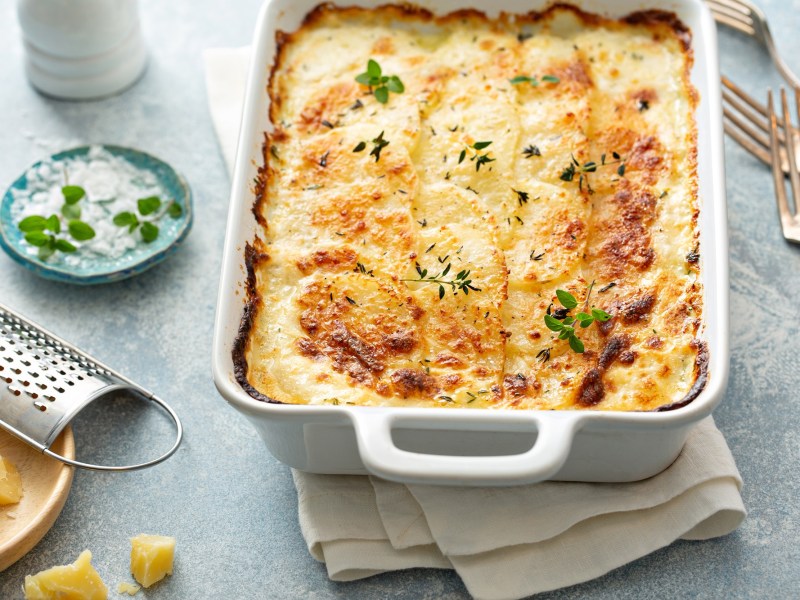 Veganes Kartoffelgratin in weißer Auflaufform, mit Kräutern bestreut, drumherum einige Zutaten.