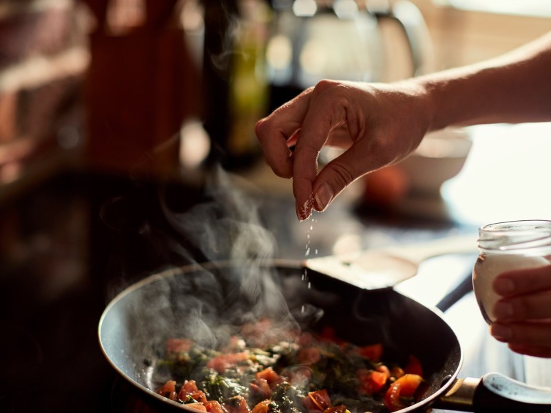 Speisen würzen: Eine Hand würzt ein Gericht in einer Pfanne.