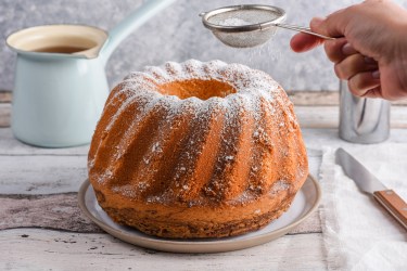Ein Amaretto-Kuchen in Gugelhupfform, der auf einem Teller steht und gerade mit Puderzucker bestäubt wird. Neben dem Kuchen liegt auf einem Stück Stoff ein Messer. Im Hintergrund steht ein gefülltes Gefäß.