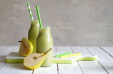 Zwei kleine Glasflaschen mit hellgrünem Birnen-Feldsalat-Smoothie, die auf einem weißen Holzbrett stehen. Auf dem Brett liegen außerdem ein gelber und ein grüner Strohhalm und es liegen 2 Birnenhälften mit im Bild. In den Glasflaschen steckt auch jeweils ein grüner Strohhalm.