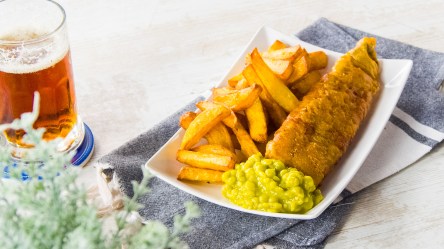 Fish and Chips in einer Schale, auf Zeitungspapier angerichtet. Dabei Erbsenpüree und Zitrone.