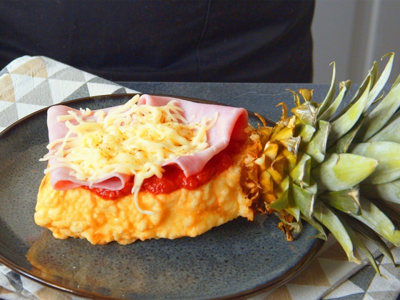 Ein Stück Ananas-Pizza, das aus einem mit Hefeteig ummantelten und frittierten Ananasstück besteht, welches mit Tomatensoße Schinken und Käse belegt ist. Der Strunk ist noch am Ananasstück dran. Die Ananas-Pizza liegt auf einem dunkelgrauen Teller, der auf einem gemusterten Küchentuch liegt.