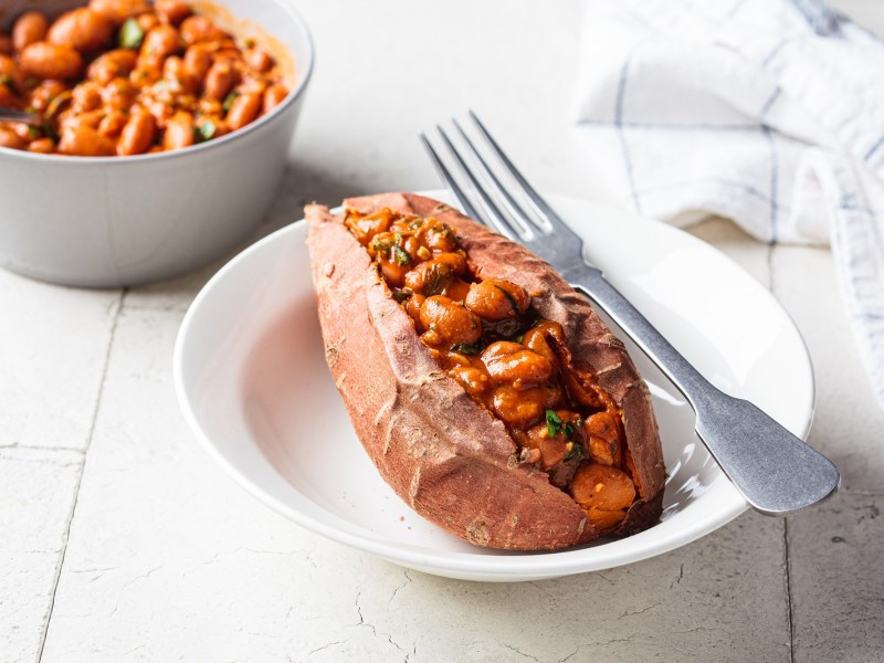 Eine gefüllte Süßkartoffel mit Bohnen auf einem weißen Teller. Im Hintergrund steht eine Schüssel Bohnen.