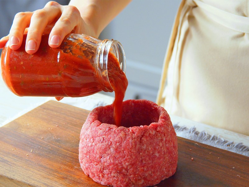 Ein roher Hackfleisch-Cup, der gerade mit passierten Tomaten beziehungsweise Tomatensoße aufgefüllt wird. Der Hackfleisch-Cup steht auf einem Holzbrett. Außerdem ist eine Hand und der Unterkörper der Köchin zu sehen.